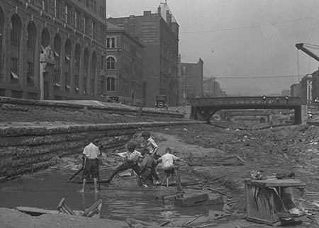 A photo of the construction of Cincinnati's unfinished subway system showing children playing in what was the Cincinnati canal.  There are building and a bridge in the background.  The date of the photograph is June 10, 1920.